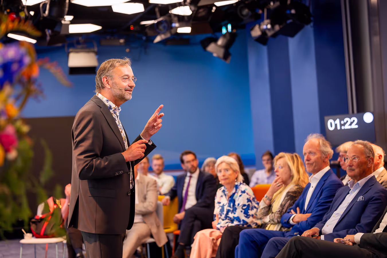 Spreker in een zakelijk pak presenteert voor een zittend publiek tijdens een evenement, met een timer op de achtergrond die 01:22 aangeeft.